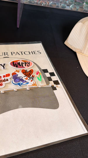 Beige trucker hat beside a "Pick Your Patches" display of colorful iron-on patches and a pearl chain on a black table — DIY hat-decorating station.