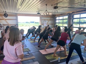 Group yoga class in a sunlit barn-style studio with open garage doors overlooking green fields, participants on mats practicing a forward lunge stretch in an indoor-outdoor community workout.