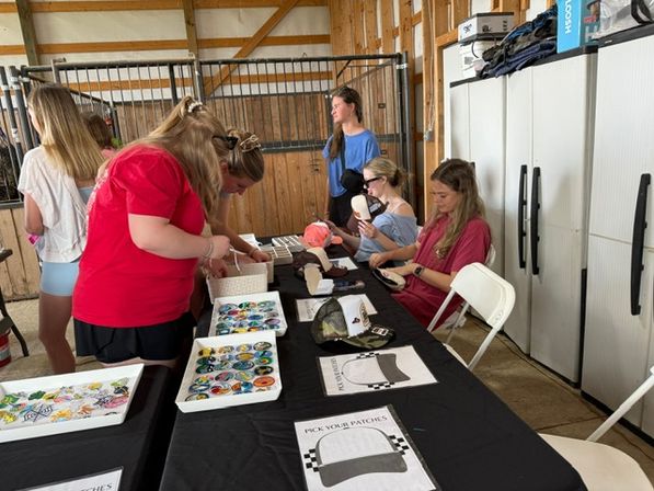 Visitors at a DIY patch-making station inside a wooden barn stable, browsing colorful round patches, hat templates, and craft supplies on a long black-covered table.