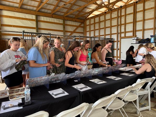 Young women browsing patches and accessories at a long vendor table inside a sunlit wooden barn market, with black tablecloth, clear display bins and folding chairs.