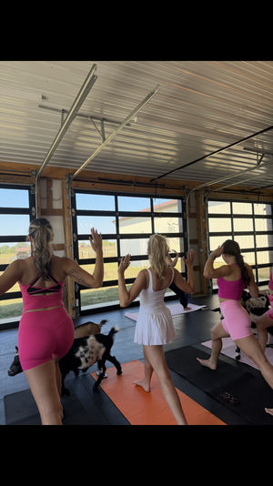 Group of women practicing goat yoga on colorful mats in a bright barn-style studio with glass garage doors and a rural farm view