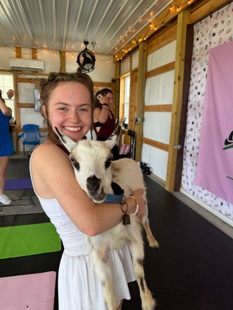Smiling young woman holding a small white-and-black goat in a bright barn-style indoor goat yoga class with yoga mats and string lights visible