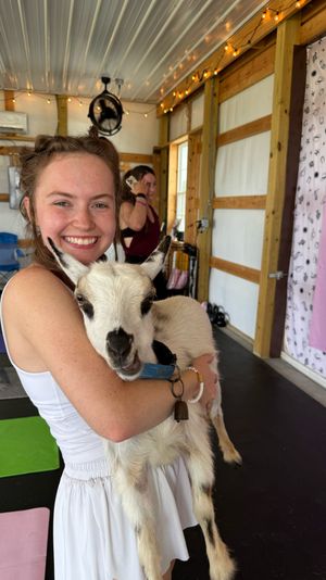 Smiling young woman holding a small white-and-black goat in a bright barn-style indoor goat yoga class with yoga mats and string lights visible
