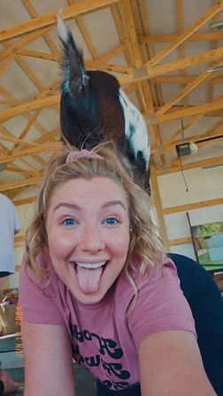 Playful selfie of a woman sticking her tongue out while a small spotted goat stands on her back under a wooden-beam barn ceiling