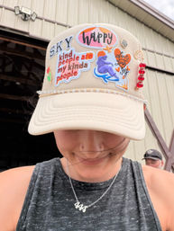 Beige trucker hat decked with colorful embroidered patches (happy, SKY, kind people are my kinda people), pearl trim and red-bead dangle, worn by a smiling person looking down in front of a metal barn-like building.
