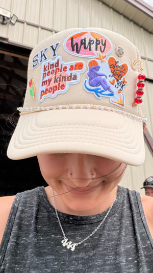 Beige trucker hat decked with colorful embroidered patches (happy, SKY, kind people are my kinda people), pearl trim and red-bead dangle, worn by a smiling person looking down in front of a metal barn-like building.