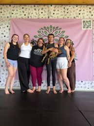 Six barefoot women smiling in an indoor studio in front of a pink banner with a green tree logo, one woman holding a small brown goat — playful goat yoga group photo