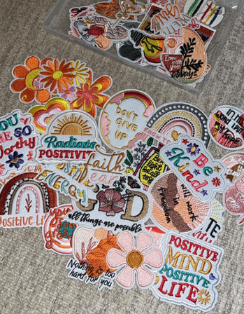 Colorful embroidered patches with positive affirmations, rainbows, suns and flowers scattered on a gray carpet and in a clear tray — DIY craft supply image.