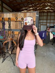 Person in lavender activewear adjusts a decorative "Be Happy" trucker hat inside an equestrian tack room with saddles, bridles, wooden stalls, and barn rafters.