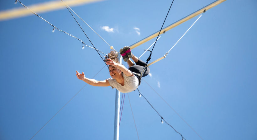 Smiling aerial performer on a trapeze at an outdoor festival, mid-swing against a clear blue sky with visible rigging and safety harness