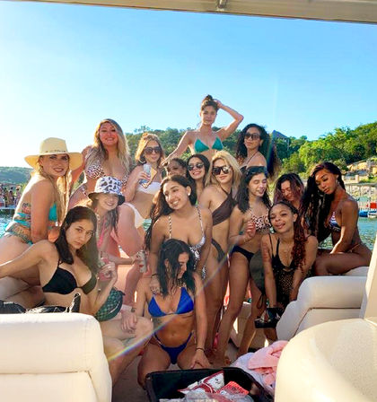 Group of women in colorful bikinis posing and holding drinks on a sunny boat during a lakeside summer party with green shoreline in the background