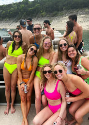 Group of friends in bright neon bikinis smiling and posing on a pontoon boat near a rocky lakeshore with tree-covered hills, enjoying a sunny summer day with canned drinks.