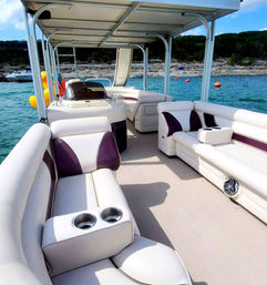 Pontoon boat interior with white and purple cushioned seating, built-in cup holders and speakers, shaded canopy over turquoise lake near rocky shoreline and orange buoys