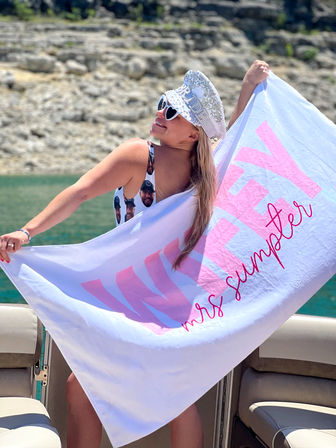 Smiling woman in heart sunglasses and a sparkly bridal captain hat holding a pink-lettered personalized towel aboard a boat on a turquoise lake with a rocky shoreline — bachelorette summer vibe