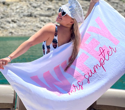 Smiling woman in heart sunglasses and a sparkly bridal captain hat holding a pink-lettered personalized towel aboard a boat on a turquoise lake with a rocky shoreline — bachelorette summer vibe