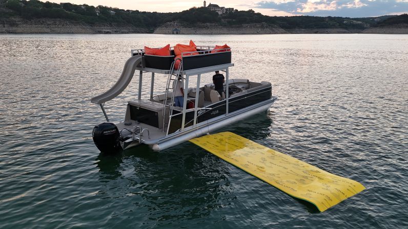 Pontoon boat with an upper-deck slide and orange cushions tethered to a long yellow water mat, floating on a calm lake at sunset with rocky shoreline in the distance
