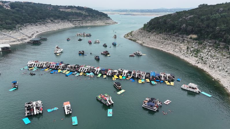 Aerial view of a bustling lake cove with dozens of pontoon boats in a long flotilla, colorful inflatable slides and floating mats, swimmers and scattered boats between rocky, tree-lined shores.