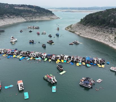 Aerial view of a bustling lake cove with dozens of pontoon boats in a long flotilla, colorful inflatable slides and floating mats, swimmers and scattered boats between rocky, tree-lined shores.