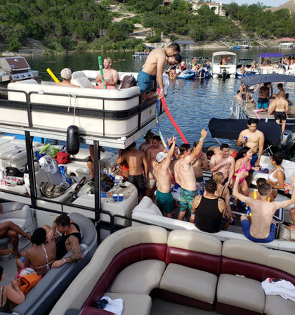 Crowded summer lake party on stacked pontoon boats with young adults dancing, drinking, and socializing between boats, pool noodles and inflatables on a sunny day.