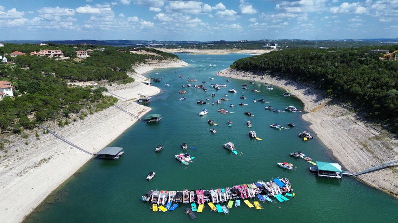 Aerial view of a busy summer lake channel with dozens of boats, colorful floats, and clustered docks between rocky, tree-lined shorelines under a sunny blue sky.