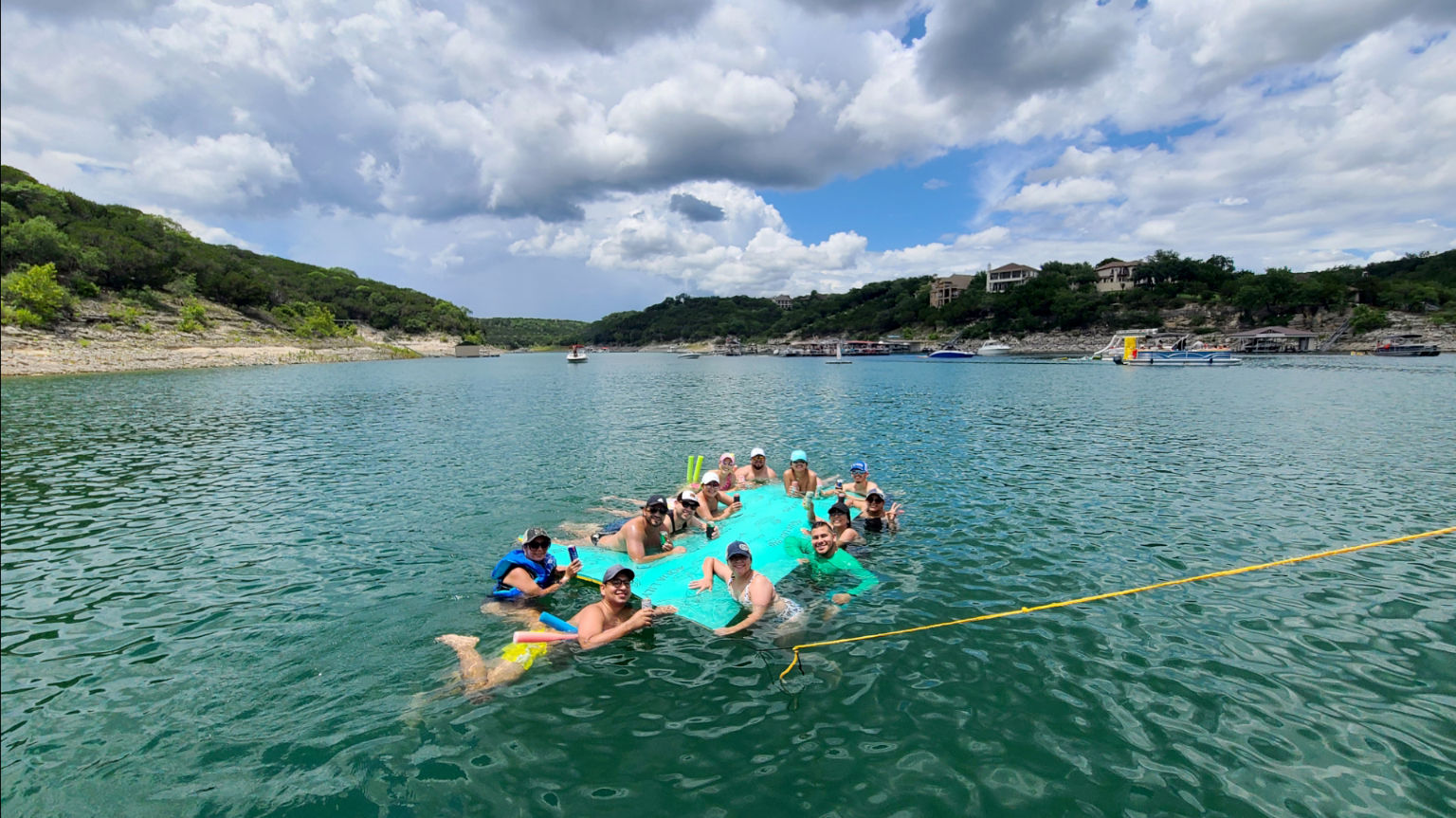 Group of friends on a bright teal inflatable raft floating in a scenic hill-lined lake cove with boats and hilltop homes beneath a dramatic partly cloudy sky
