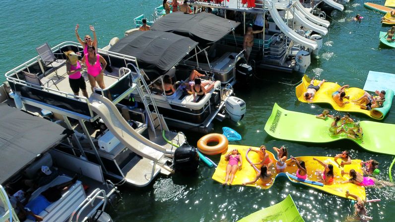 Sun-soaked lake party with multiple pontoon boats and waterslides, people lounging on bright inflatable mats and tubes in the water