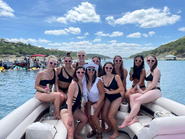 Group of friends in black swimsuits and pink heart-shaped sunglasses posing and holding canned drinks on a pontoon boat during a sunny summer lake day with tree-lined shores and other boats in the background.