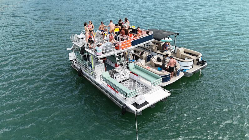 Aerial view of a multi-level pontoon party on a green lake, people lounging, sunbathing, and socializing on connected decks and cushioned seats, summer boating scene.