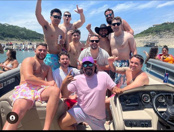 Pontoon boat summer party: group of friends in swim trunks cheering with drinks on a sunny lake, shoreline and boats in the background.