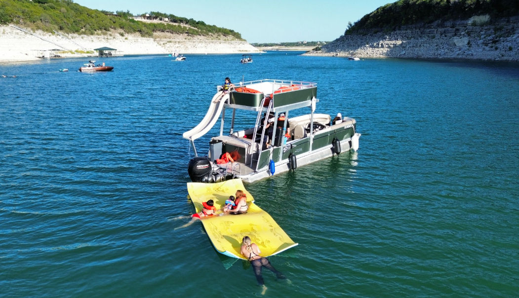 Pontoon boat with top-deck slide and yellow inflatable mat carrying swimmers and kids on a cliff-lined lake on a sunny summer day