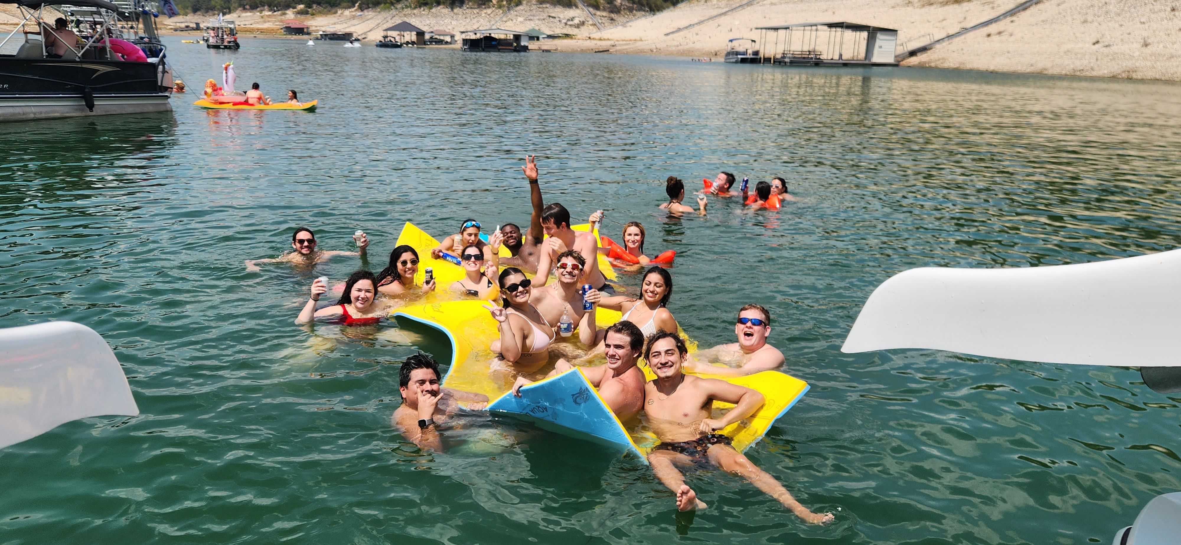 Smiling group of people lounging on a bright yellow inflatable mat in green lake water, holding drinks and posing while boats, kayaks and a sandy shoreline with docks sit in the background on a sunny summer day
