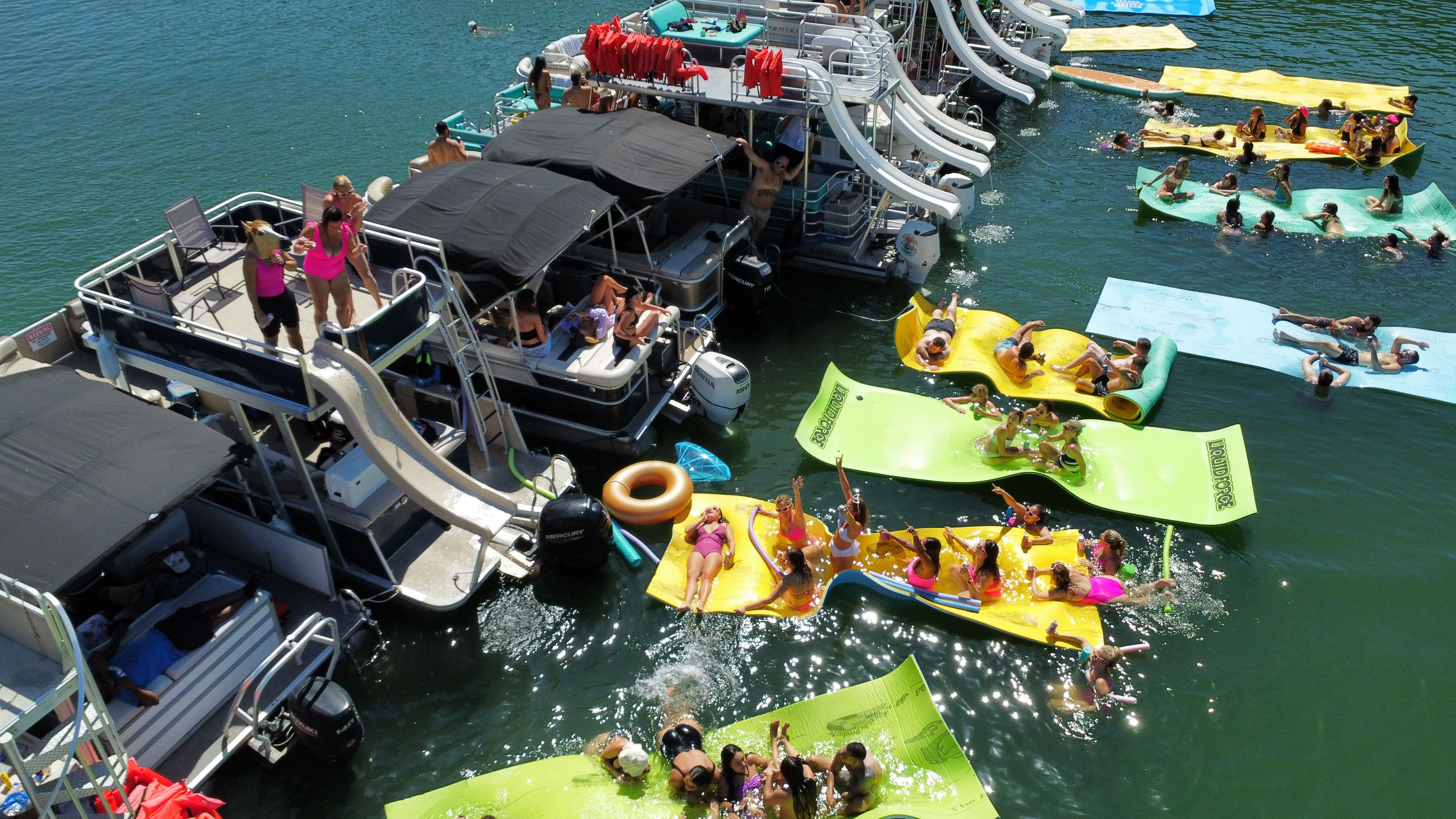 Aerial view of a sun-drenched lake party with pontoon boats and slides docked together, colorful floating mats and inflatables, and groups swimming, sunbathing, and playing on the water.