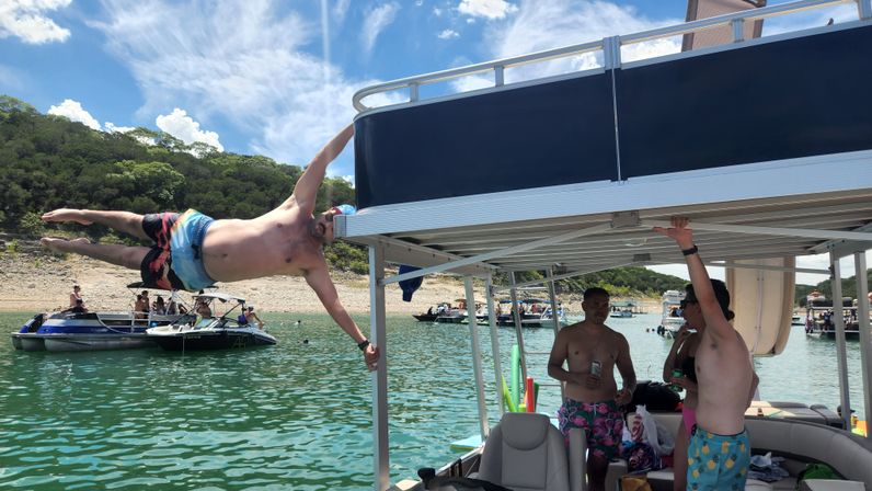 Man holding a horizontal 'human flag' from a pontoon boat canopy while friends in swim trunks relax aboard, turquoise lake with other boats and a tree-lined shore under a blue summer sky