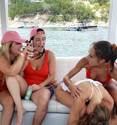 Four friends laughing on a pontoon boat at a lake, wearing swimsuits and pink caps, one holding a canned drink while another playfully leans over a friend, tree-lined shoreline and another boat in the background.