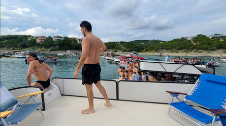 Summer pontoon boat party at a scenic lake cove — people in swimsuits on crowded boats, wooded shoreline and blue cloudy sky