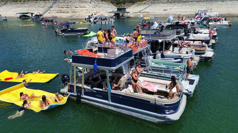 Pontoon boats tied together at a lively lake party with people sunbathing, swimming, and relaxing on bright yellow floating mats near a sandy shoreline