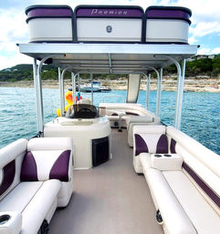 Pontoon boat interior with white and purple lounge seating, overhead canopy and slide, parked on a blue lake near a rocky shoreline — summer boating scene