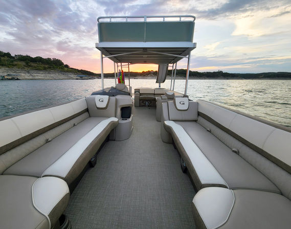 Spacious pontoon boat interior with beige bench seating, upper deck and slide, colorful pool noodles, cruising on a lake at sunset with rocky shoreline in the background.