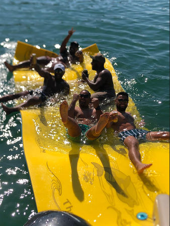 Six men relaxing and splashing on a bright yellow inflatable raft in clear blue water, wearing swim trunks and sunglasses and holding canned drinks on a sunny day.