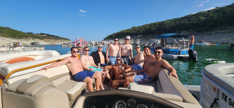 Group of friends lounging on a pontoon boat at a sunny summer lake cove, surrounded by boats, inflatables, and a rocky shoreline.
