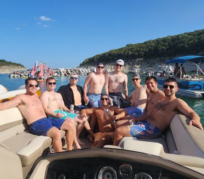 Group of friends lounging on a pontoon boat at a sunny summer lake cove, surrounded by boats, inflatables, and a rocky shoreline.