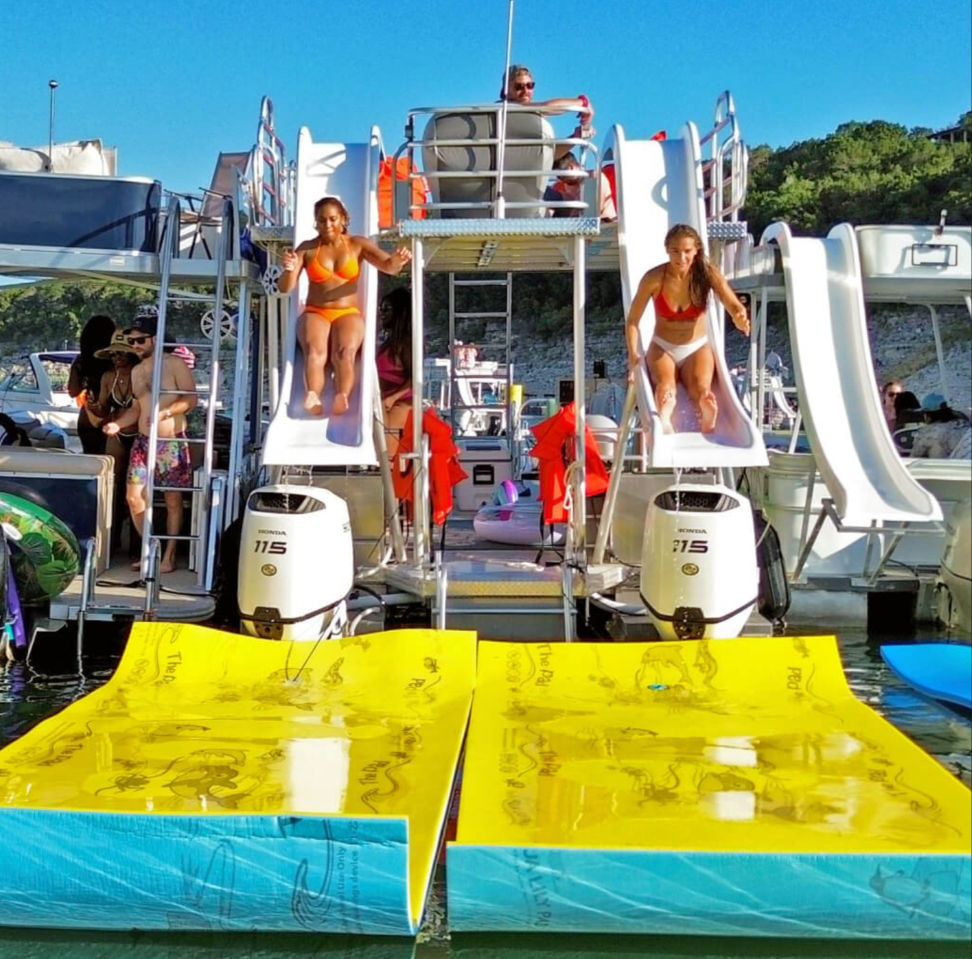 Two women in bikinis sliding down boat-mounted waterslides onto bright yellow floating mats at a lively summer lake boat party with pontoons and outboard engines under a clear blue sky