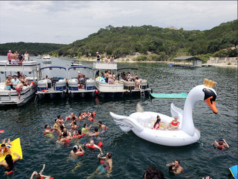 Summer lake party with several pontoon boats tied together, dozens of people swimming with red floaties and a giant inflatable swan near a tree-lined shoreline
