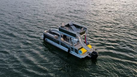 Pontoon boat with upper-deck slide, sun loungers and colorful inflatable tube floating on rippled lake water at dusk.