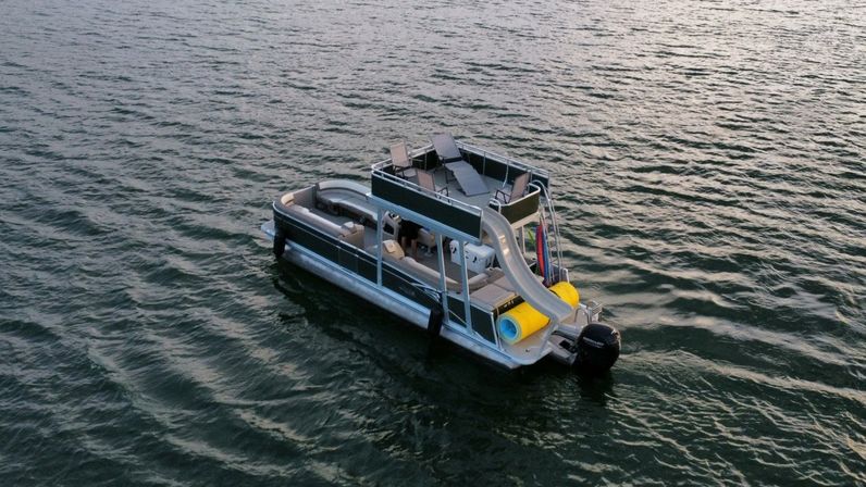 Pontoon boat with upper-deck slide, sun loungers and colorful inflatable tube floating on rippled lake water at dusk.