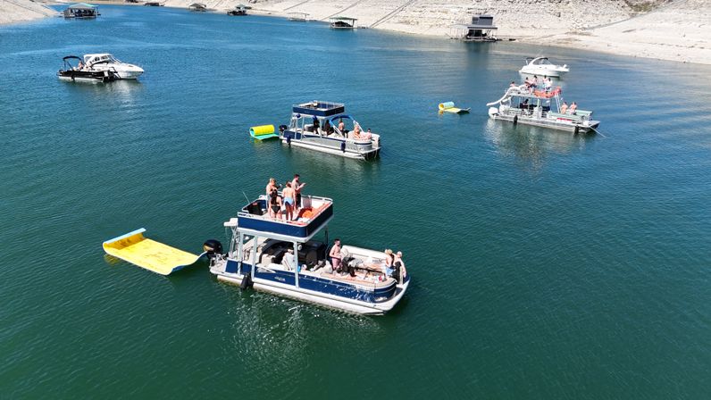 Aerial view of pontoon and motor boats anchored on a sunny lake near a rocky shoreline, groups enjoying summer boating with a yellow inflatable water ramp and floating toys.