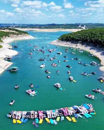 Aerial view of a crowded summer lake party with dozens of boats and colorful floating platforms anchored in turquoise water between rocky, tree-lined shores under a bright blue sky