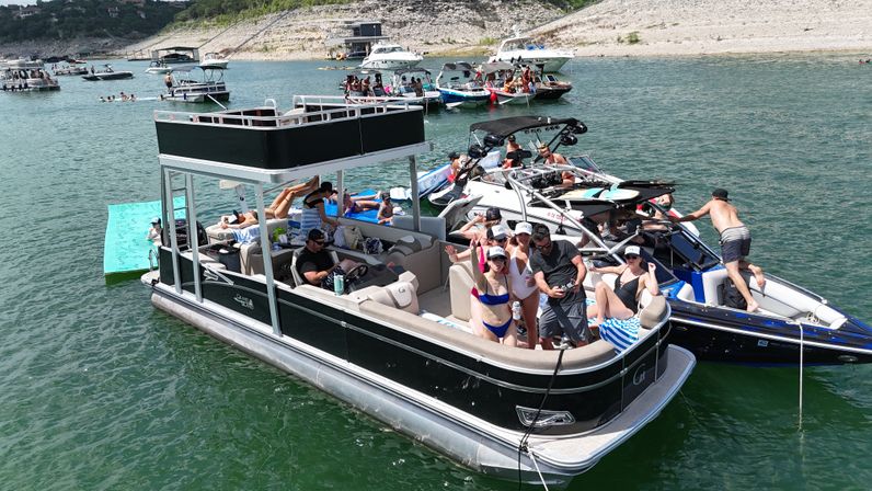 Sun-soaked lake party on a black double-decker pontoon boat surrounded by powerboats and swimmers near a rocky shoreline.