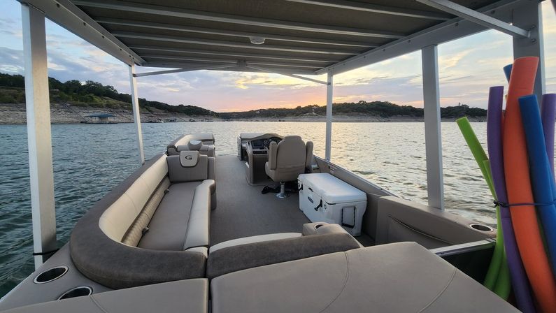 Pontoon boat interior under a canopy with curved cushioned seating, captain’s helm, white cooler and colorful pool noodles, cruising on a lake at sunset with rocky shoreline and tree-covered hills