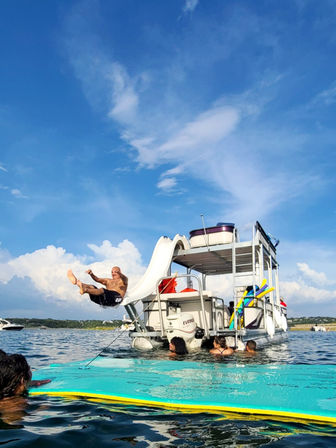 Person mid-air launching off a white slide attached to a pontoon boat into a lake, swimmers on a turquoise float nearby under a bright blue sky with puffy clouds — summer boating fun